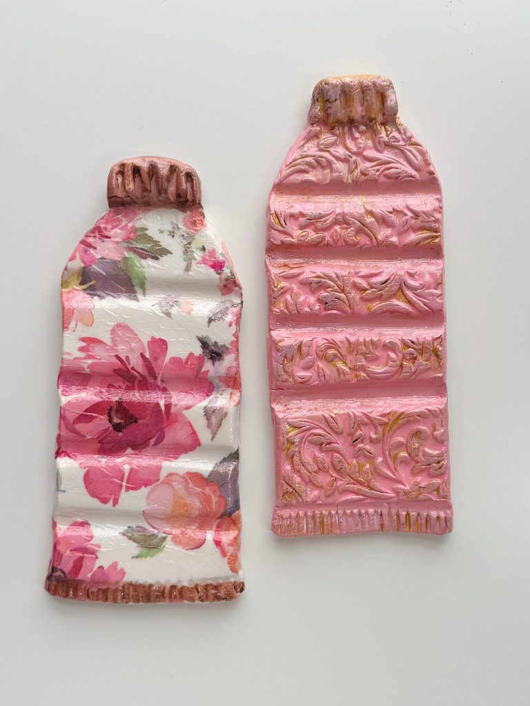 Two flat ceramic sculptures shaped like hot water bottles, one with a floral pattern and the other with pink and gold decorative designs, on a white background.