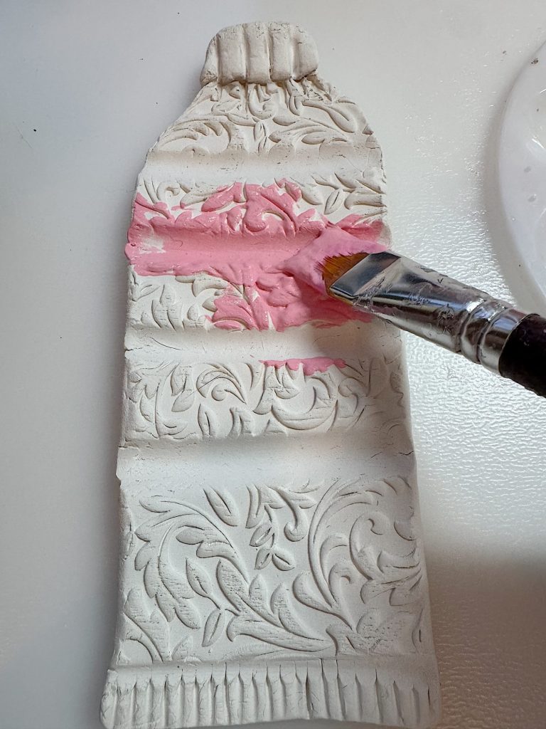 A paintbrush applies pink paint to a textured, decorative clay sculpture shaped like a paint tube lying on a white surface.