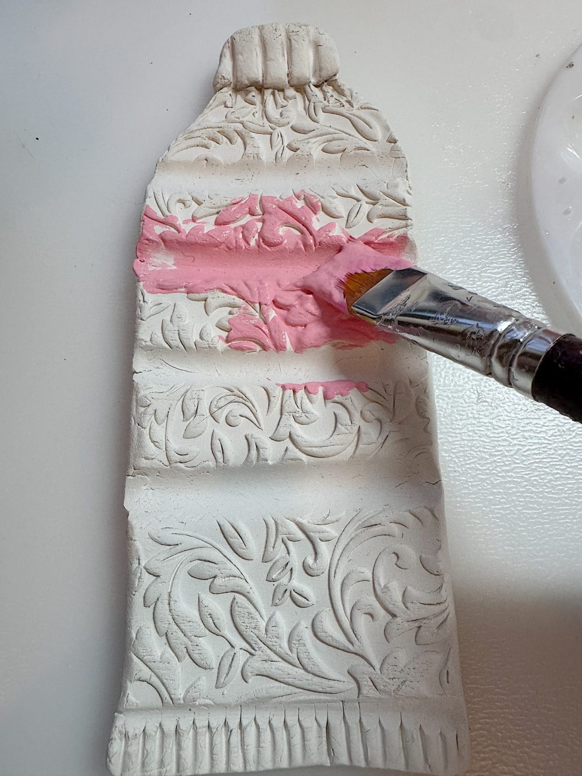 A paintbrush applies pink paint to a textured, decorative clay sculpture shaped like a paint tube lying on a white surface.