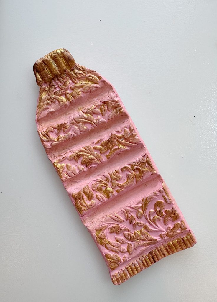 A sculpted object shaped like a flattened tube of paint, decorated with pink and gold ornate patterns, placed on a white surface.