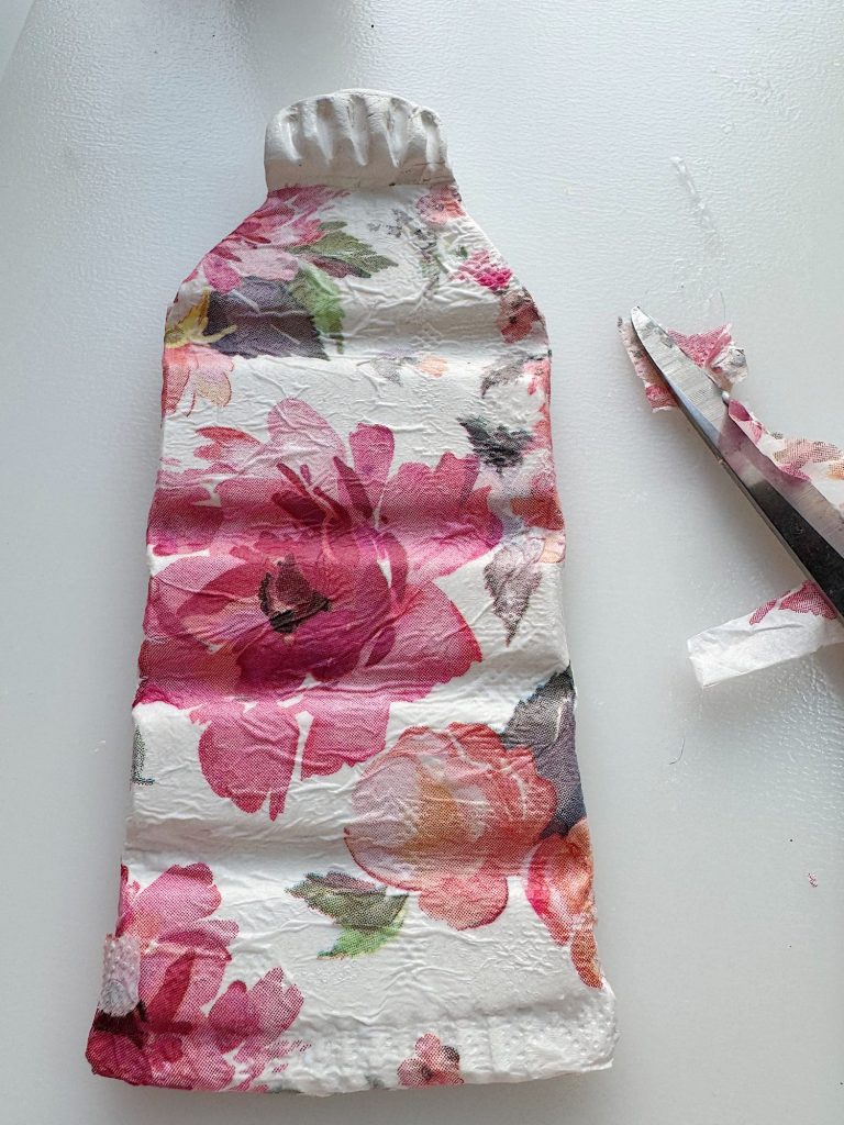 A floral fabric cover, shaped to fit a hot water bottle, lies on a white surface next to a pair of scissors and some trimmed fabric scraps.