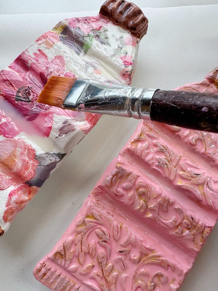 A paintbrush rests on two pieces of textured, ornate material, one decorated with floral patterns and the other painted pink with raised designs.