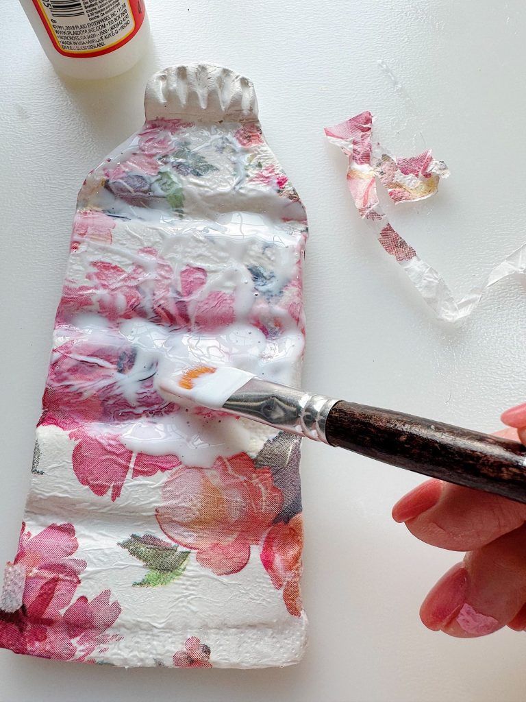 A hand uses a paintbrush to apply glue onto a floral-patterned piece of paper attached to a flattened plastic bottle.