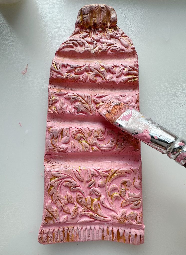 A textured sculpture shaped like a paint tube, decorated with floral patterns, is being painted pink with a brush.