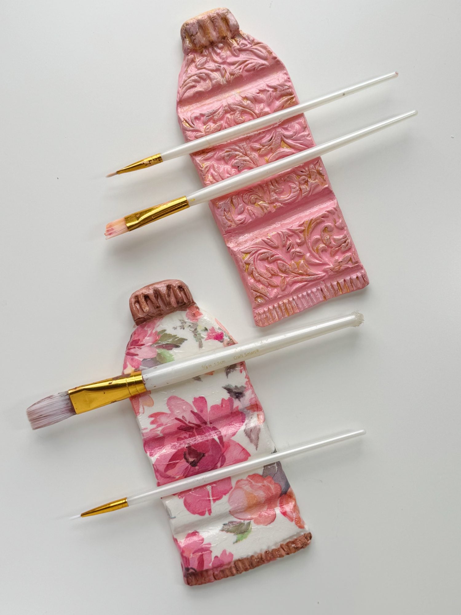 Two pink ceramic paint tube sculptures, one with floral patterns and one with ornate designs, are placed on a white surface with three paintbrushes laid across them.