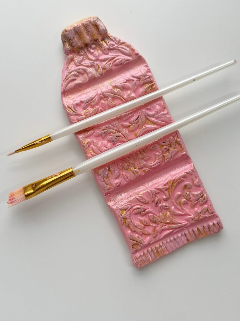 A flat, decorative pink paint tube sculpture with ornate patterns, placed beneath two white-handled paintbrushes on a white surface.