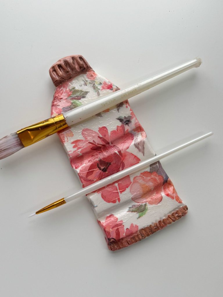 A ceramic tube painted with a floral design lies on a white surface, with two paintbrushes placed across it.