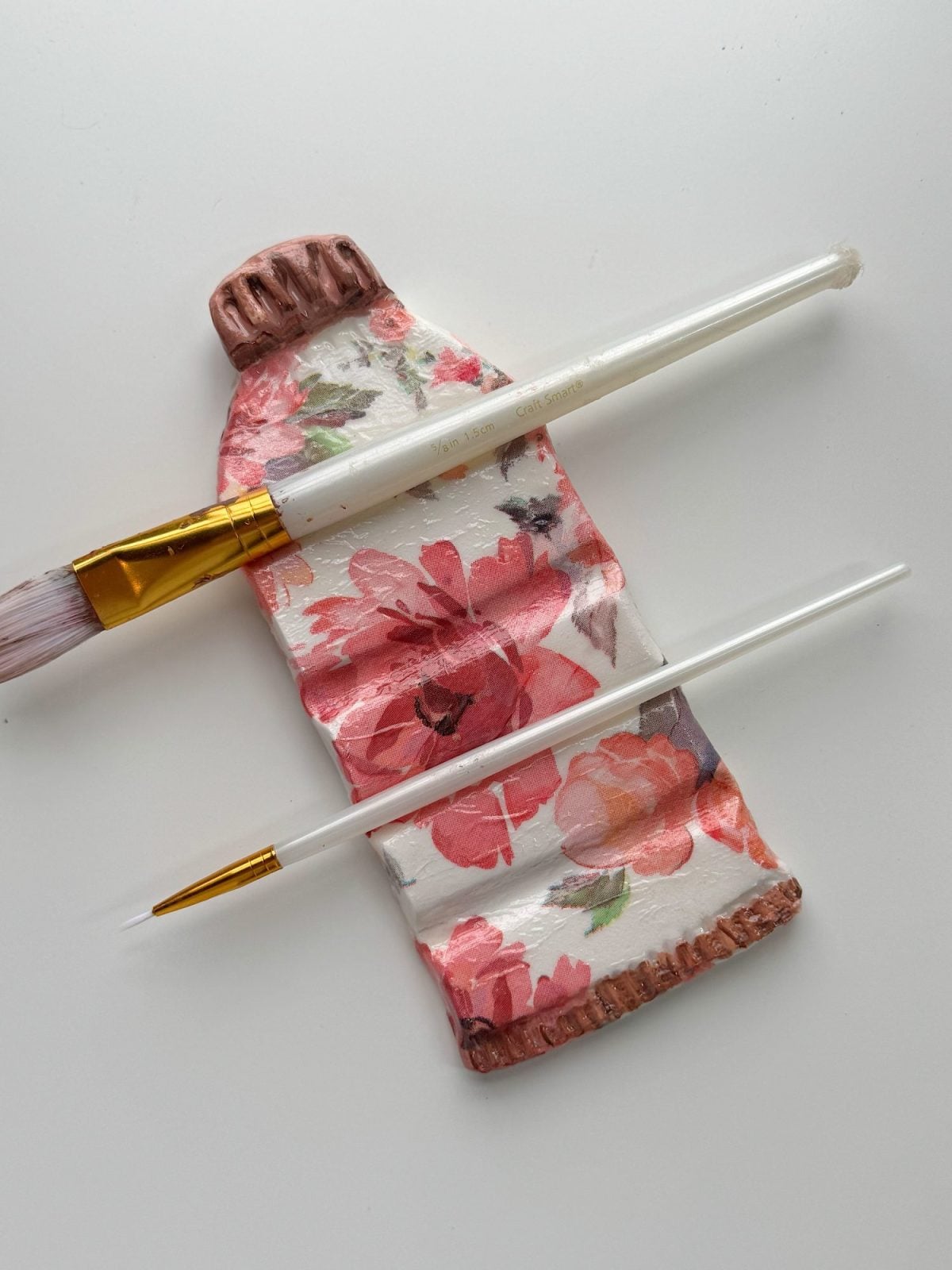 A ceramic tube painted with a floral design lies on a white surface, with two paintbrushes placed across it.