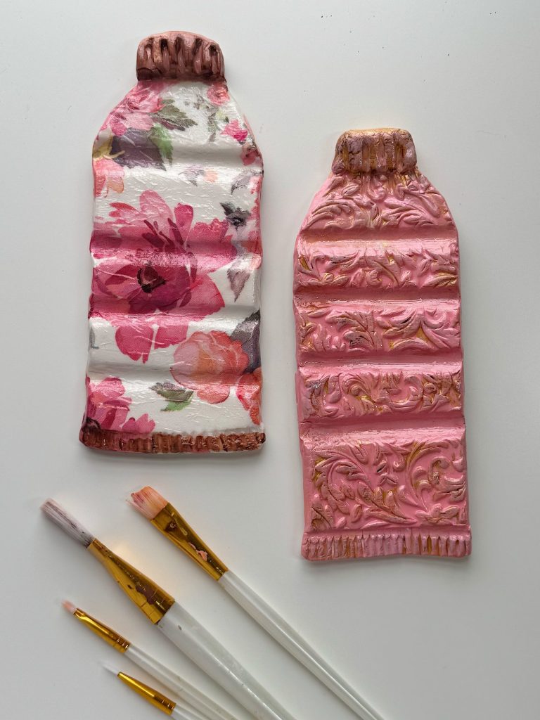Two sculpted paint tubes, one with a floral pattern and one with ornate embossed designs, are placed beside three paintbrushes on a white surface.