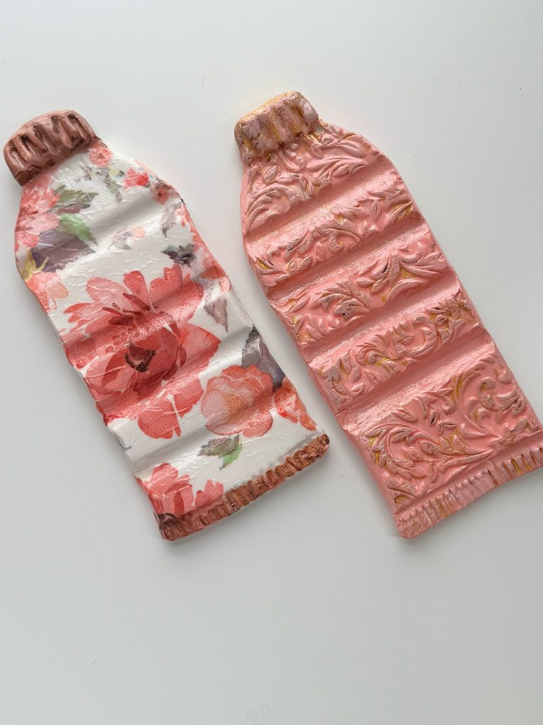 Two decorative objects shaped like flattened paint tubes, one with a red floral pattern and the other with a pink embossed design, placed on a white surface.