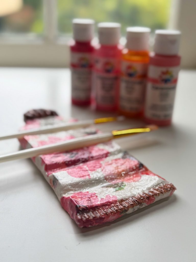 Five bottles of acrylic paint, two paintbrushes, and a rectangular object wrapped in pink floral-patterned plastic sit on a white surface near a window.