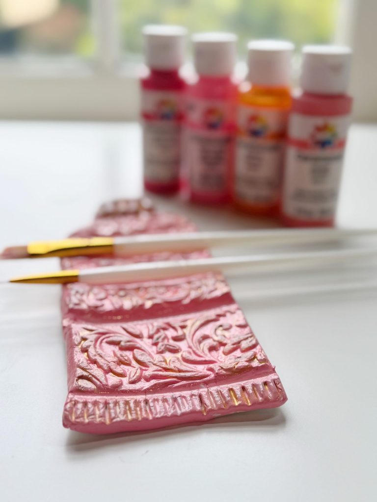 Two paintbrushes rest on a pink, ornate paintbrush holder, with four bottles of red, pink, and orange acrylic paint in the background on a white surface.