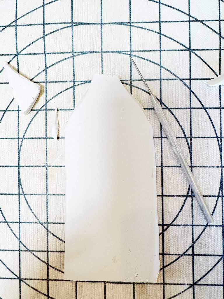 A rectangular piece of white clay with trimmed edges lies on a gridded surface next to a craft knife.