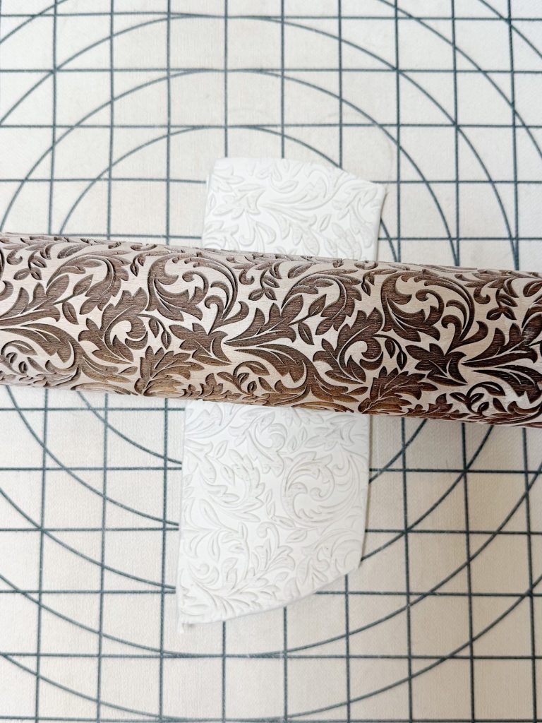 A patterned rolling pin embosses a sheet of white dough with a floral design on a gridded surface.