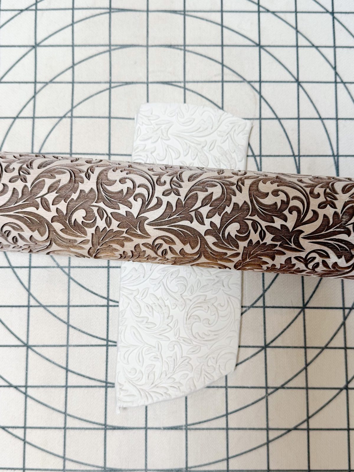 A patterned rolling pin embosses a sheet of white dough with a floral design on a gridded surface.