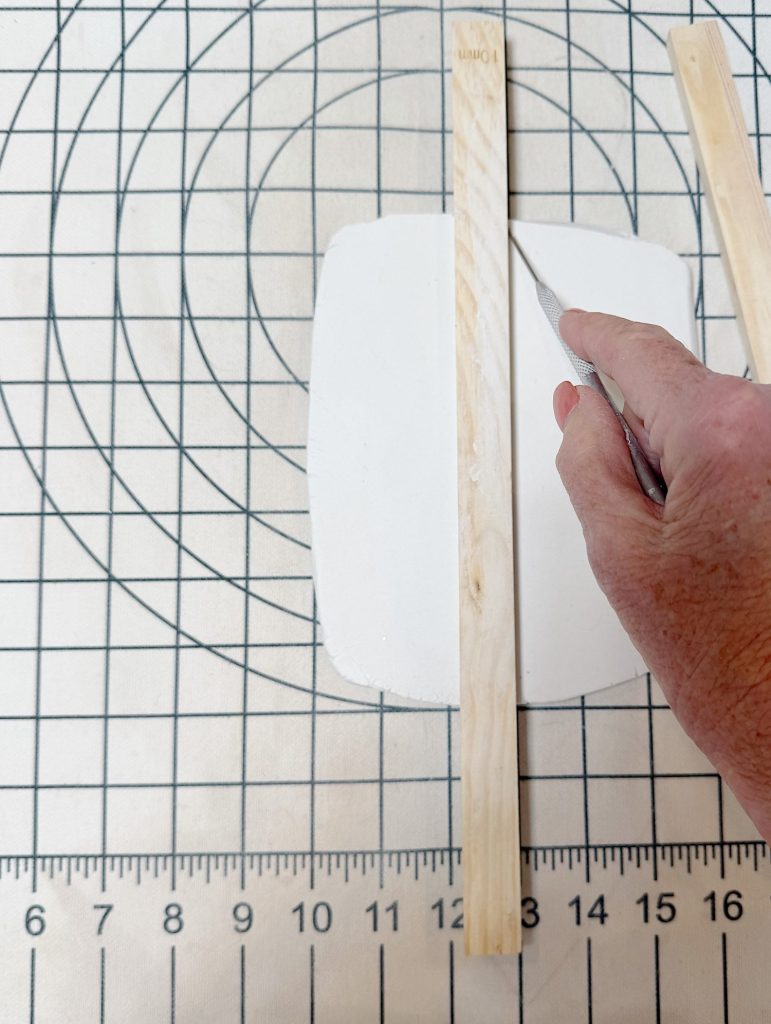 A hand uses a craft knife to cut a piece of white clay, guided by two wooden sticks, on a gridded cutting mat with a ruler and concentric circles.