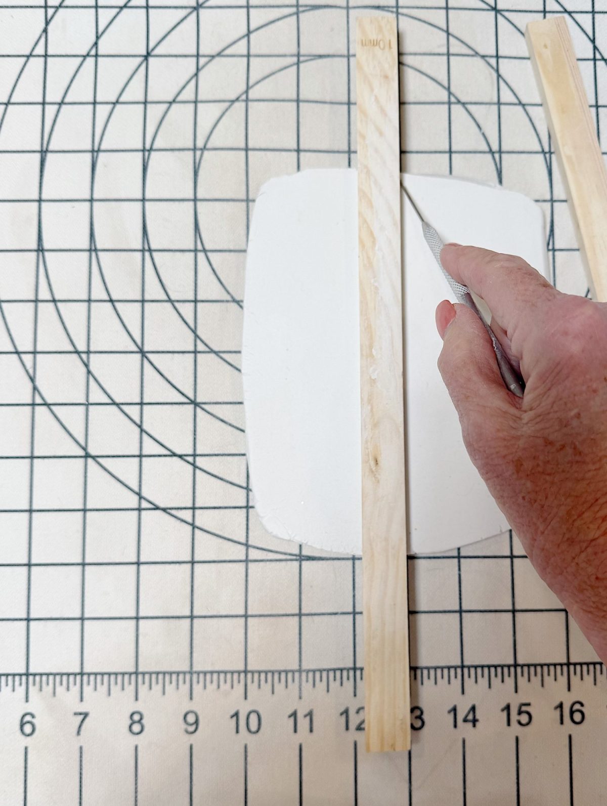 A hand uses a craft knife to cut a piece of white clay, guided by two wooden sticks, on a gridded cutting mat with a ruler and concentric circles.