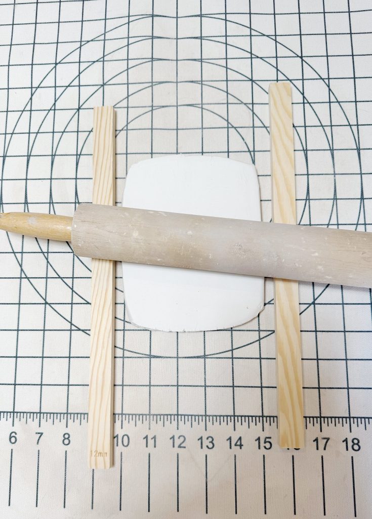 A rolling pin flattens white clay between two wooden slats on a gridded mat with measurement markings.