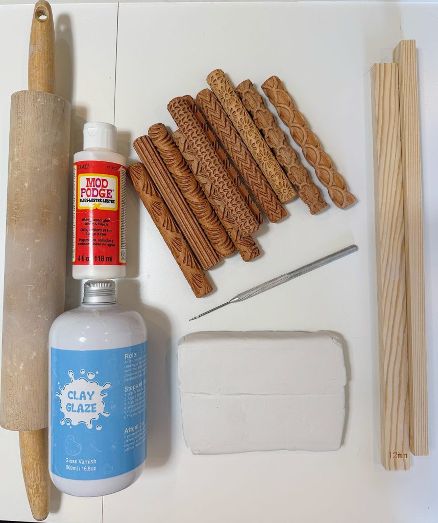 A rolling pin, textured wooden rolling tools, Mod Podge, a metal tool, wood planks, a bottle of clay glaze, and a block of modeling clay on a white surface.
