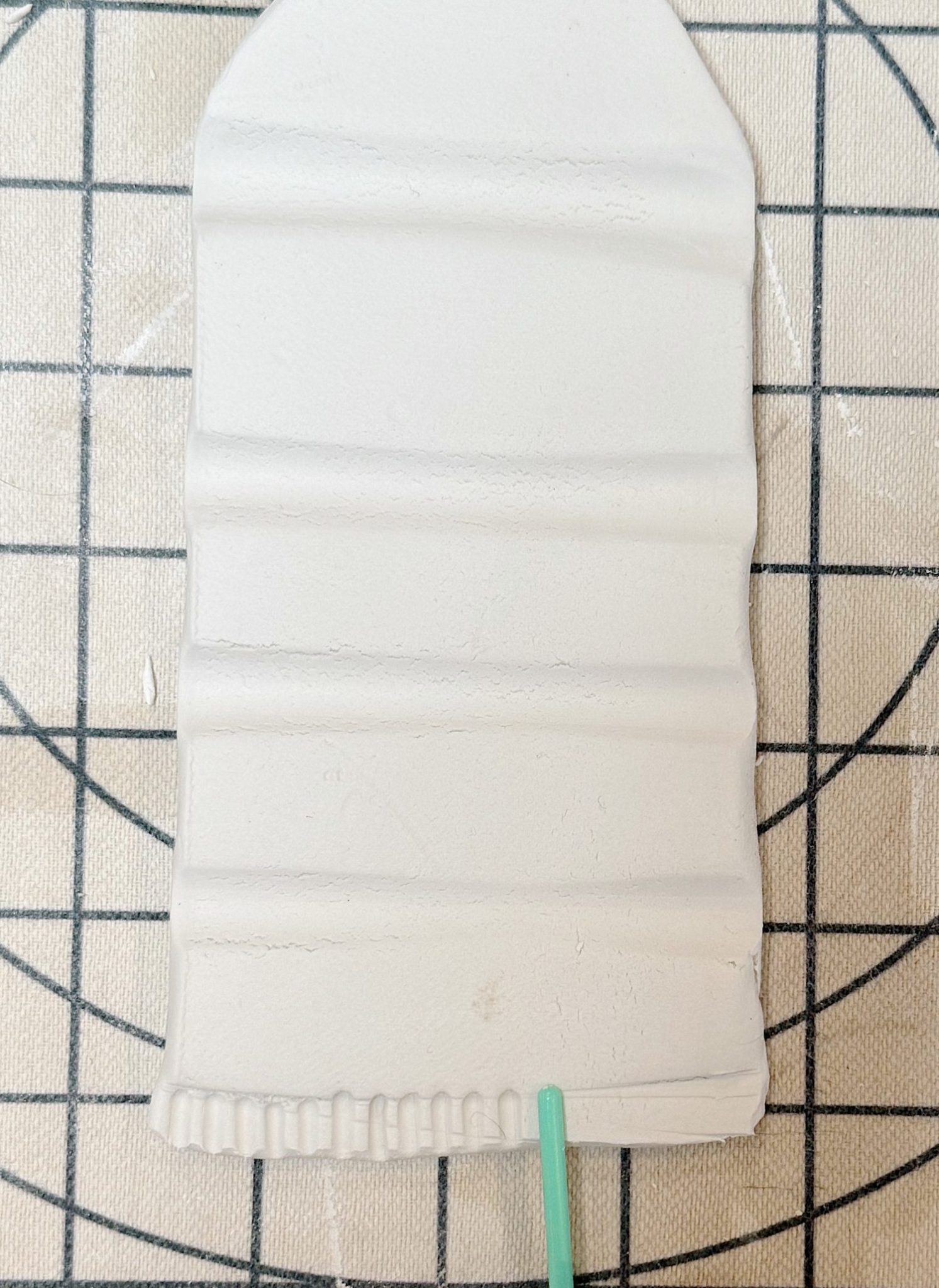 A rectangular piece of white clay with horizontal ridges lies on a grid surface; a green tool touches the bottom edge.