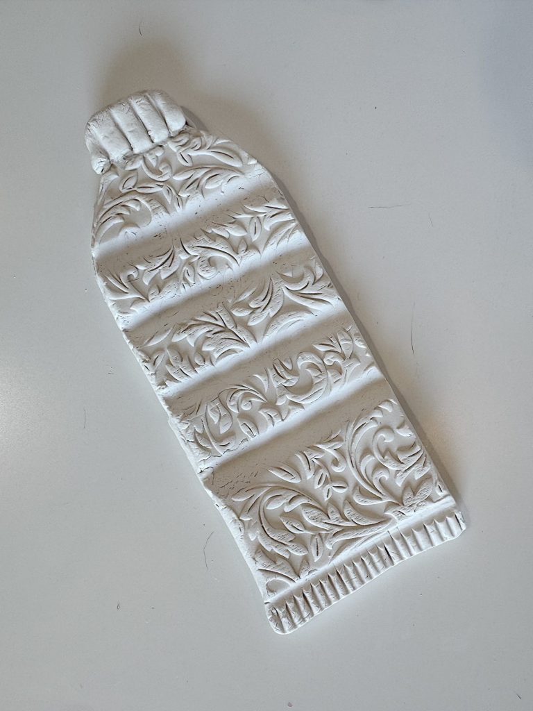 A white, decorative object shaped like a plastic water bottle features embossed floral patterns, set against a plain background.