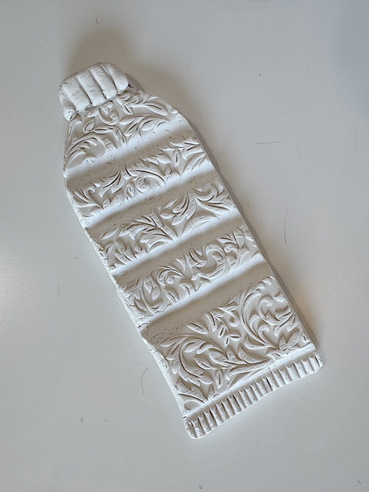 A white, decorative object shaped like a plastic water bottle features embossed floral patterns, set against a plain background.