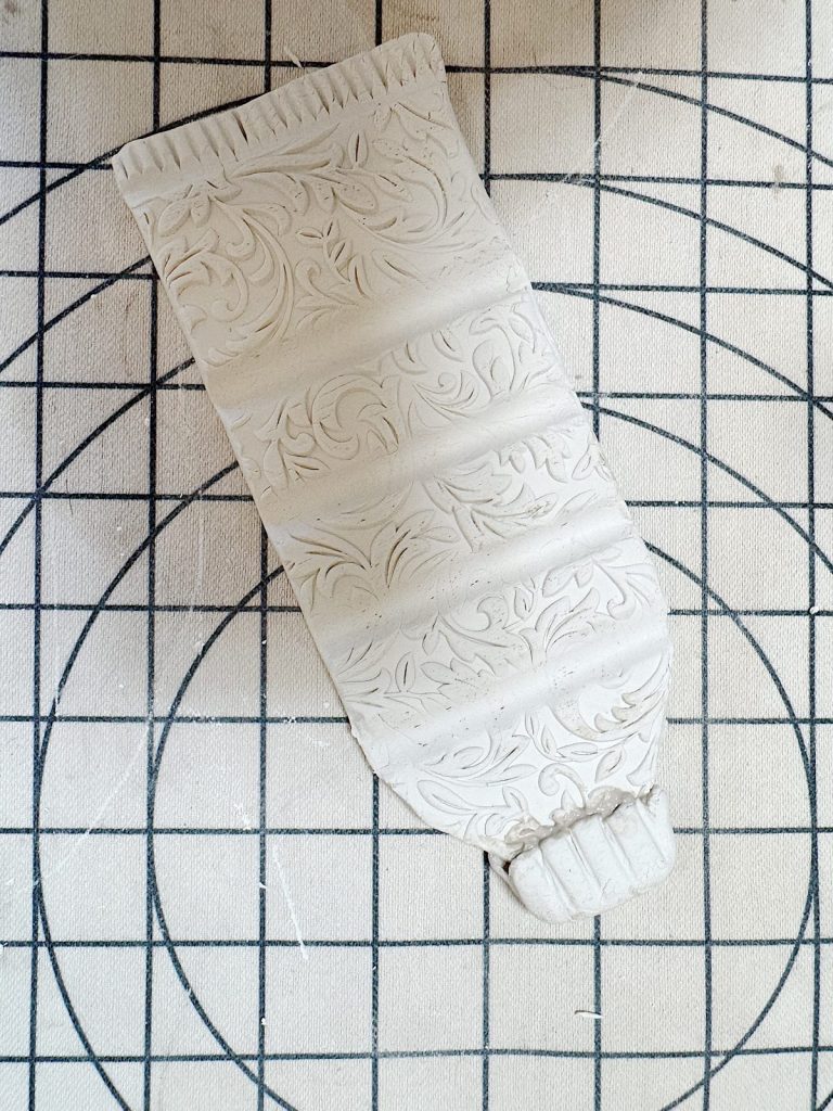 A rectangular piece of white clay with embossed floral patterns lies on a gridded surface; the clay is shaped to resemble a flattened tube.