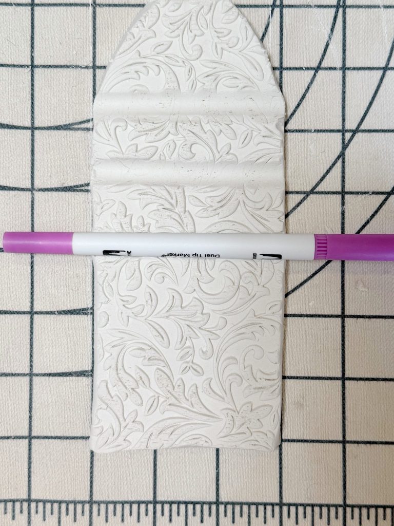 A textured piece of white clay with floral patterns lies on a grid surface, with a purple and white marker placed horizontally across it.