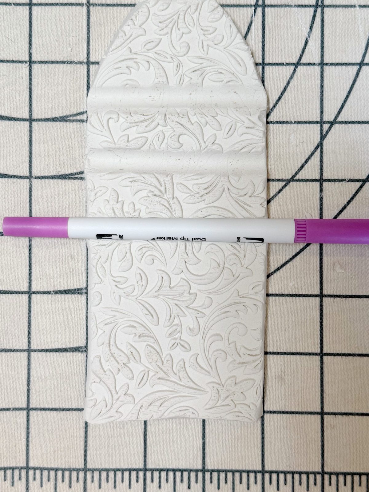 A textured piece of white clay with floral patterns lies on a grid surface, with a purple and white marker placed horizontally across it.
