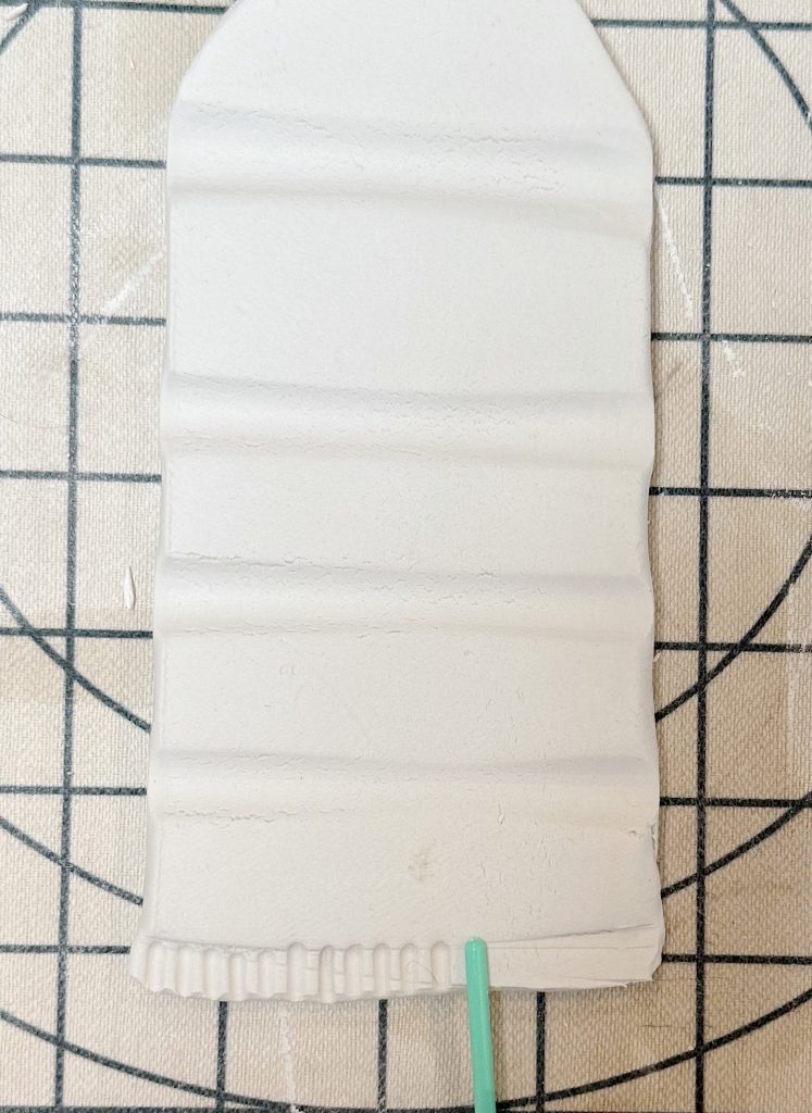 A rectangular piece of white clay with horizontal ridges lies on a grid surface; a green tool touches the bottom edge.