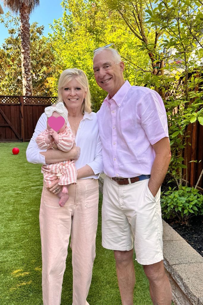 Two adults stand outside on grass, smiling at the camera; one holds a baby wearing pink stripes. Trees and a wooden fence are in the background. The baby's face is covered with a heart.