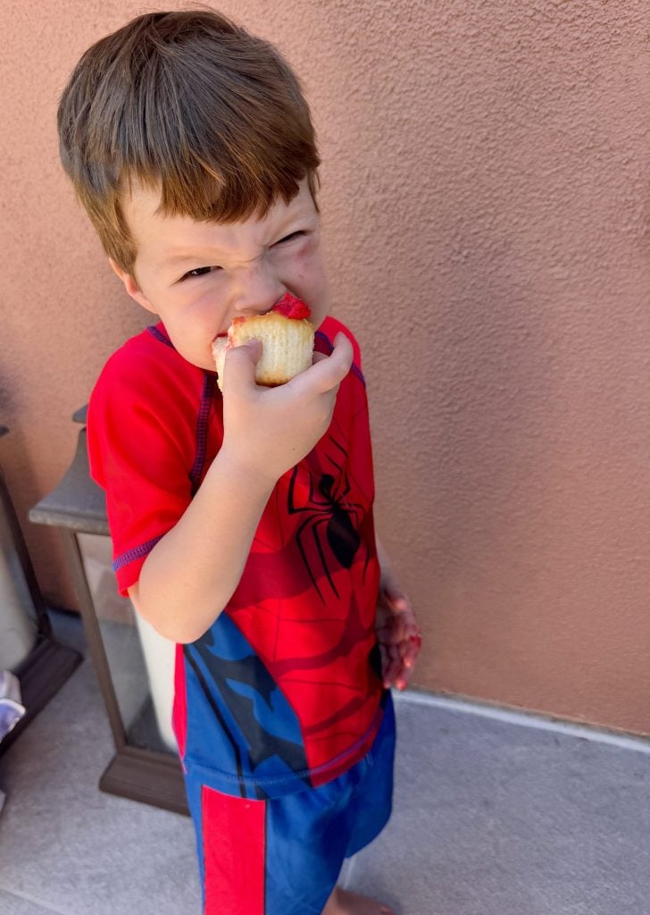 Young child in a red and blue Spider-Man outfit stands outside, squinting while eating a cupcake with pink frosting.
