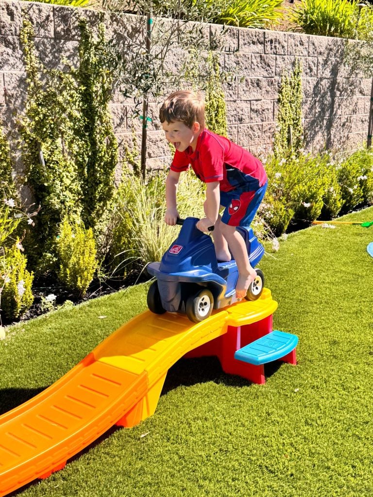 A young boy in a red outfit rides a toy car down a small plastic ramp on artificial grass in a backyard.
