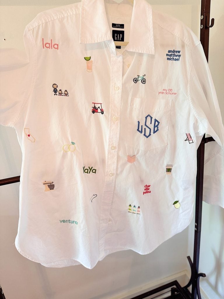A white button-down shirt with various colorful embroidered designs, including text, small figures, drinks, vehicles, and objects, displayed on a hanger.