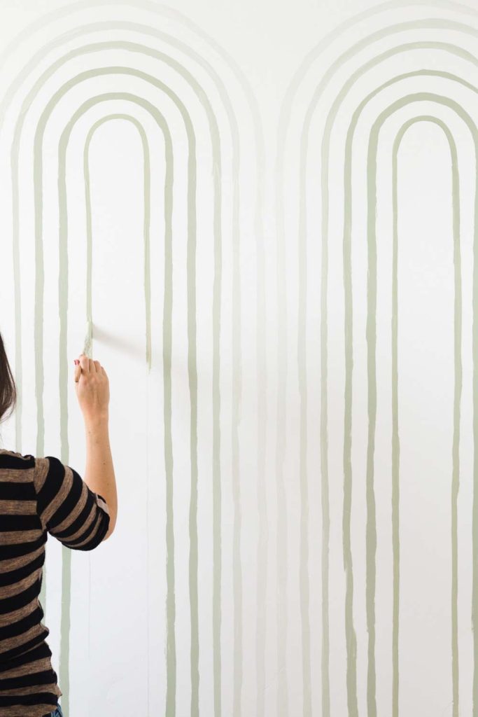 Person in striped shirt painting vertical arching lines on a white wall with a brush; only upper body and hand are visible.