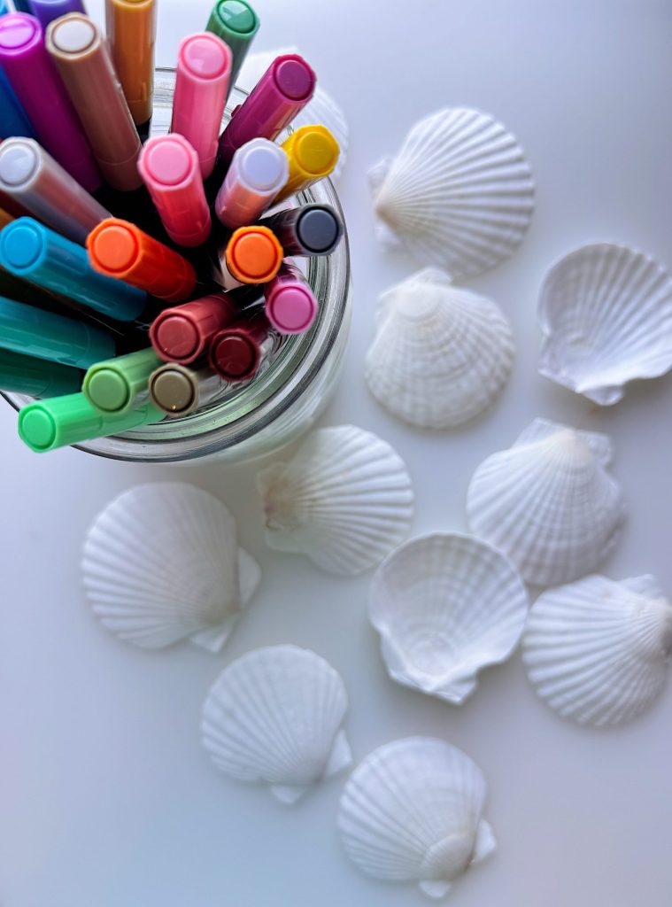 A top-down view of colorful markers in a jar next to scattered white seashells on a white surface.
