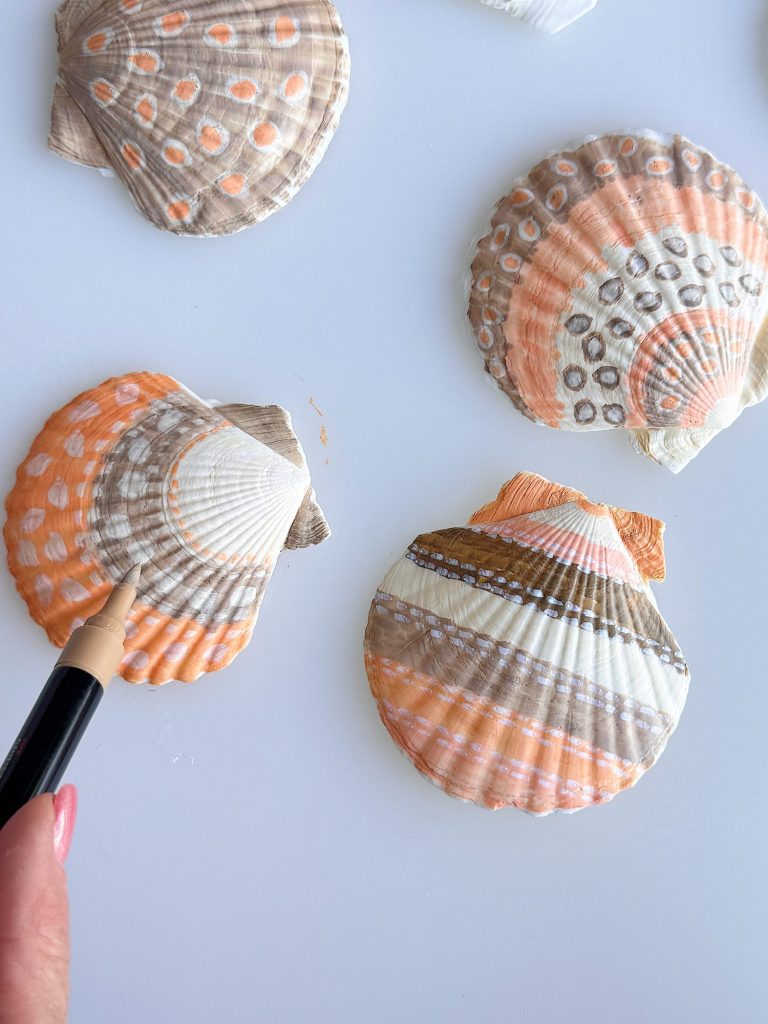 A hand uses a marker to decorate scallop seashells with geometric patterns and dots in orange, white, brown, and gray tones on a white surface.