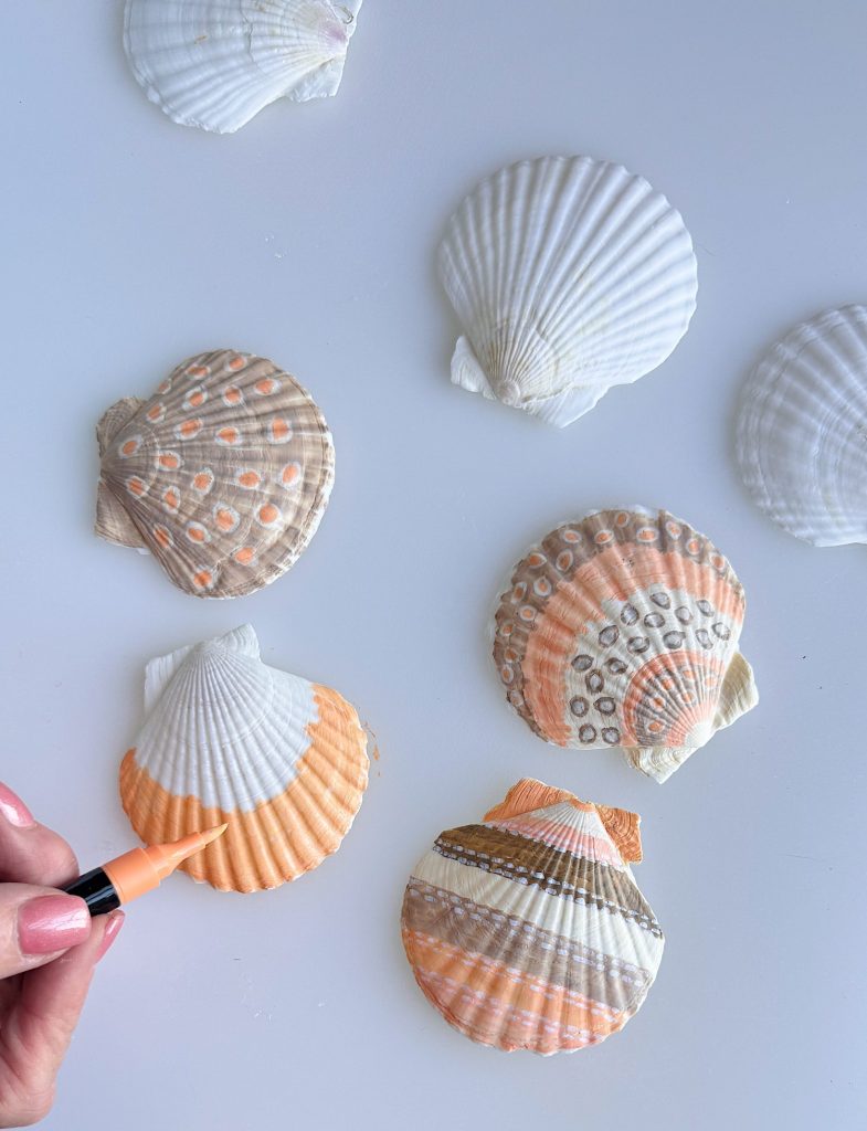 Several seashells are decorated with orange and gray patterns, while a hand uses an orange marker to color another shell on a white surface.