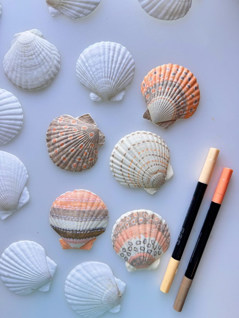 Several white and patterned seashells are arranged on a flat surface next to two dual-tip markers, one beige and one peach.