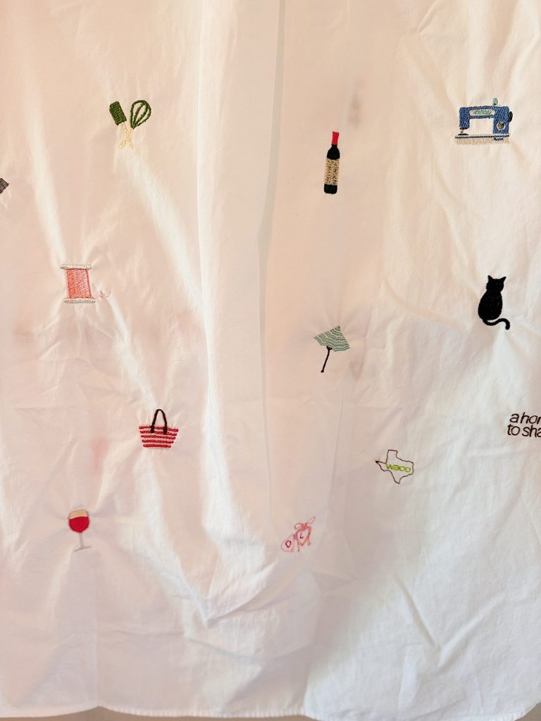 White fabric with small embroidered images including a wine bottle, sewing machine, cat, wine glass, basket, umbrella, plant, and other assorted objects scattered across the surface.