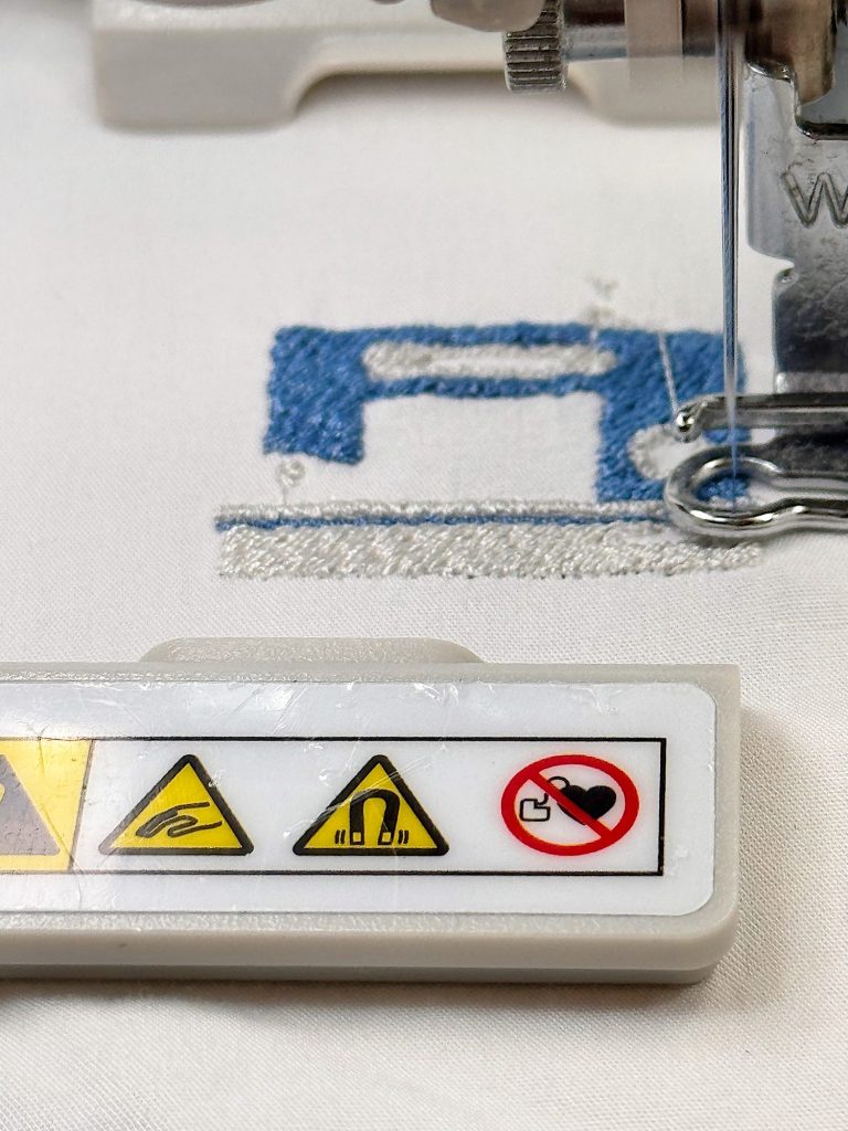 Close-up of a sewing machine needle and warning label on a plastic part, with blue thread stitching a machine icon on white fabric.