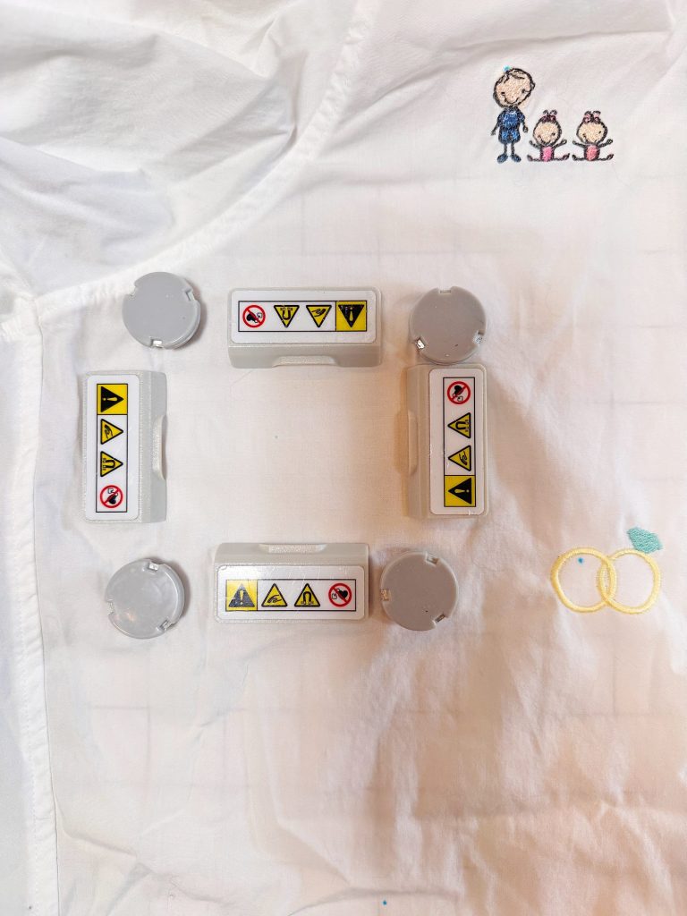 Four gray rectangular and four round plastic objects with warning labels are arranged in a square on a white cloth with a small cartoon family embroidered in the top right corner.