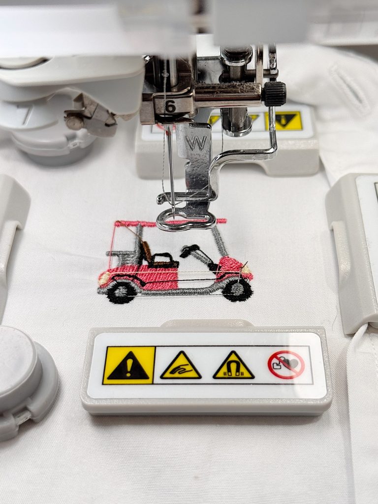 A sewing machine embroidering a pink golf cart design on white fabric, with warning label stickers visible on the machine.