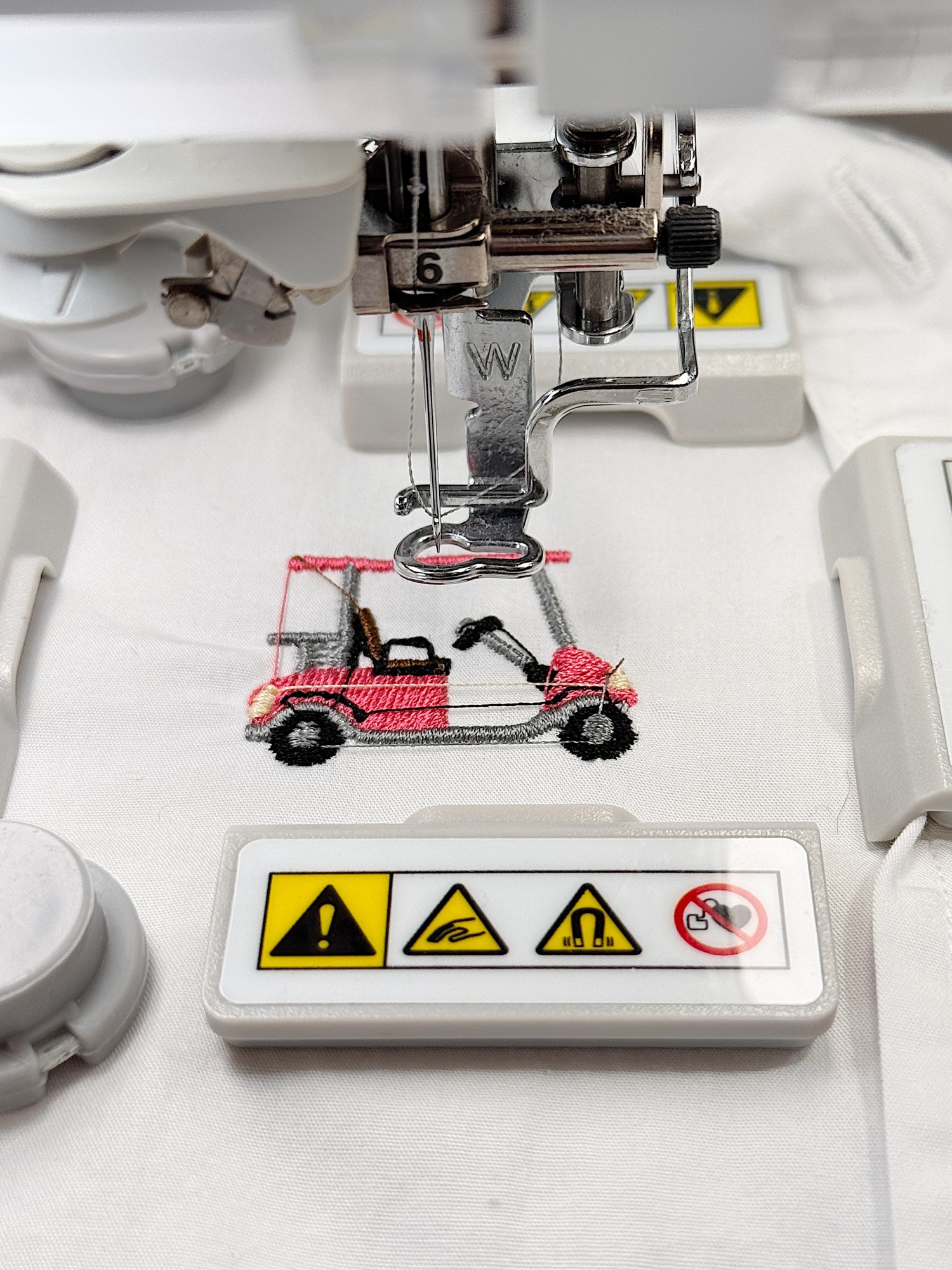 A sewing machine embroidering a pink golf cart design on white fabric, with warning label stickers visible on the machine.
