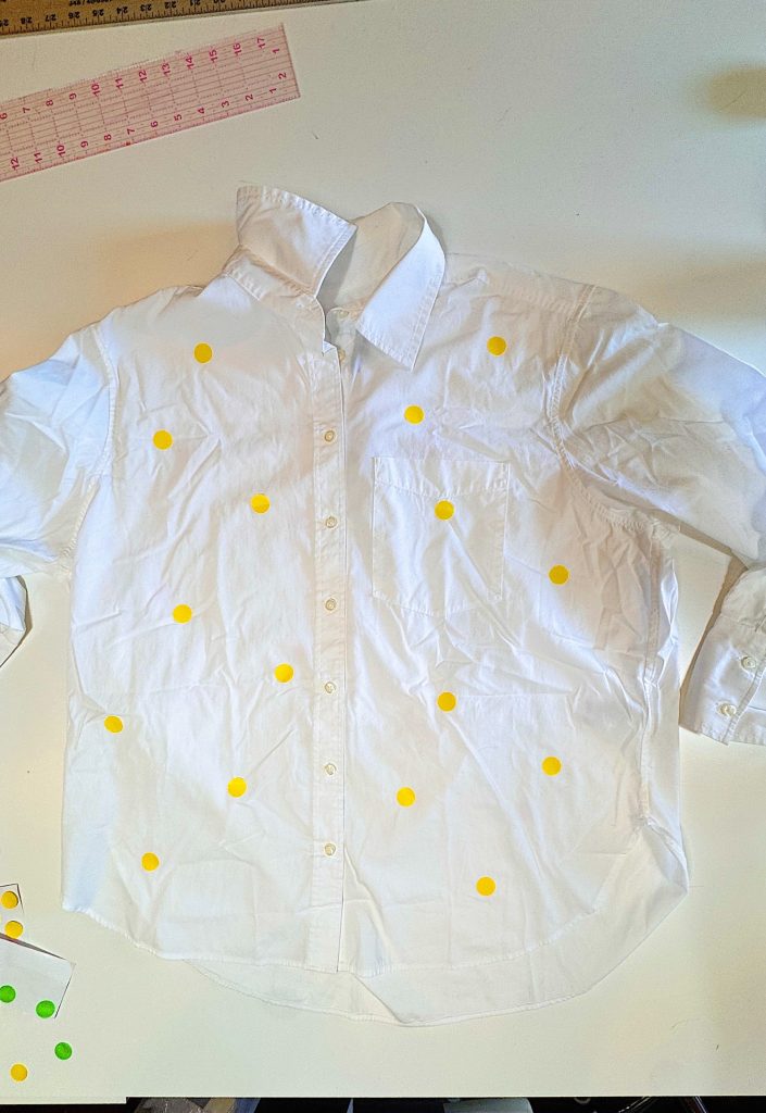 A white button-up shirt laid flat on a table, decorated with randomly placed yellow dot stickers. A ruler and more stickers are visible nearby.
