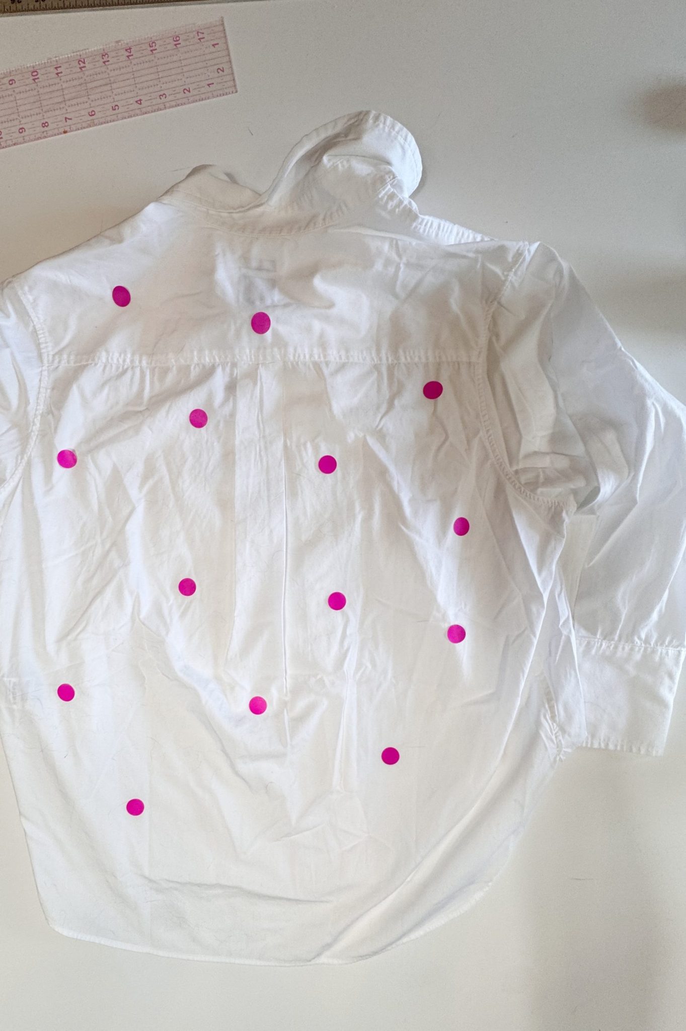 A white button-down shirt laid flat on a table, with multiple pink circular stickers placed on its back. A pink ruler is visible in the top left corner.