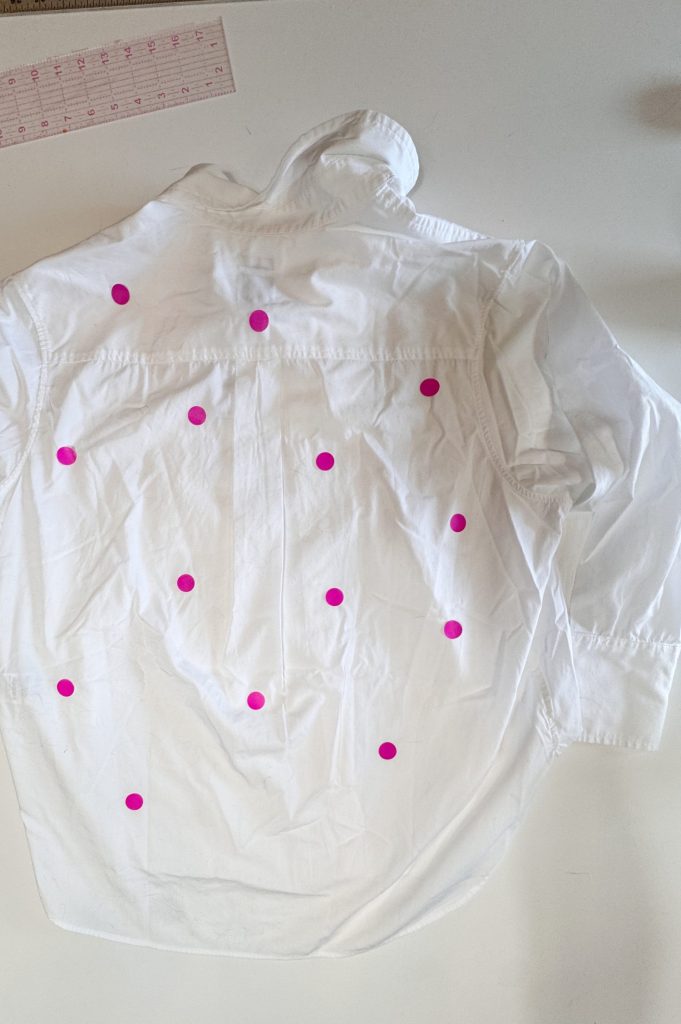 A white button-down shirt laid flat on a table, with multiple pink circular stickers placed on its back. A pink ruler is visible in the top left corner.