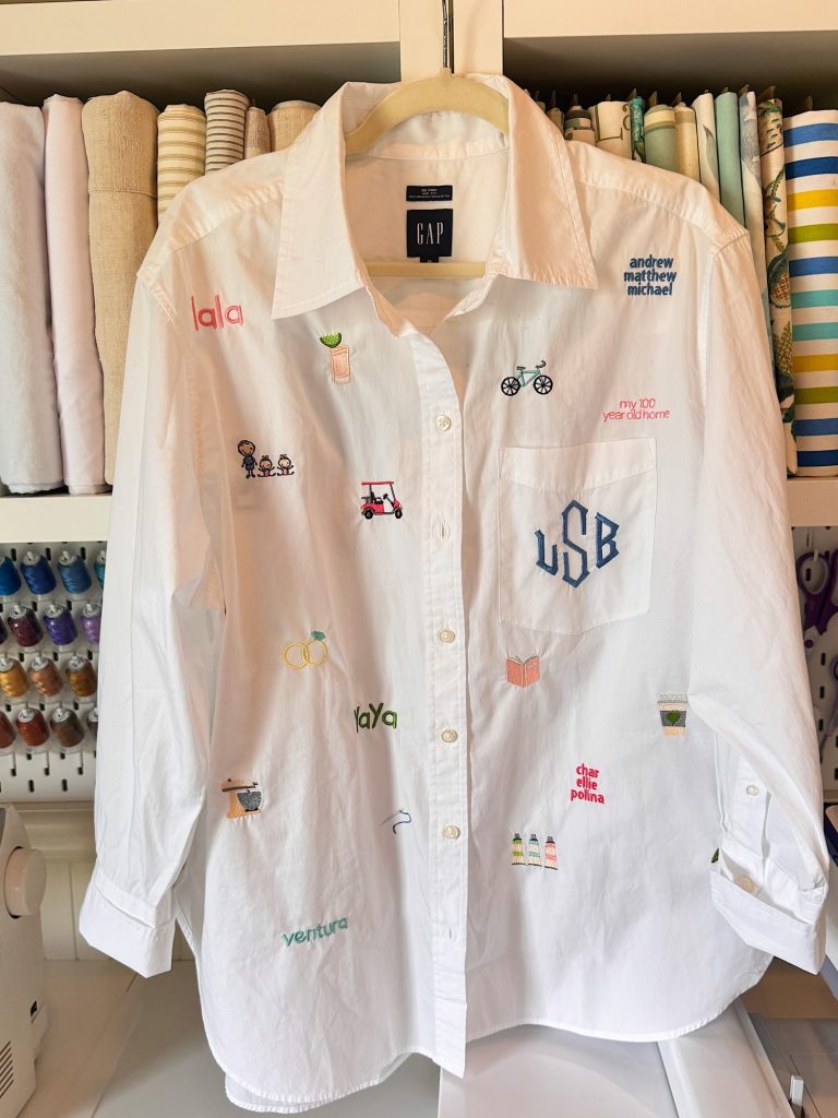 A white GAP button-up shirt hangs on a hanger, decorated with colorful embroidered words and small images. Sewing materials and fabric are visible in the background.