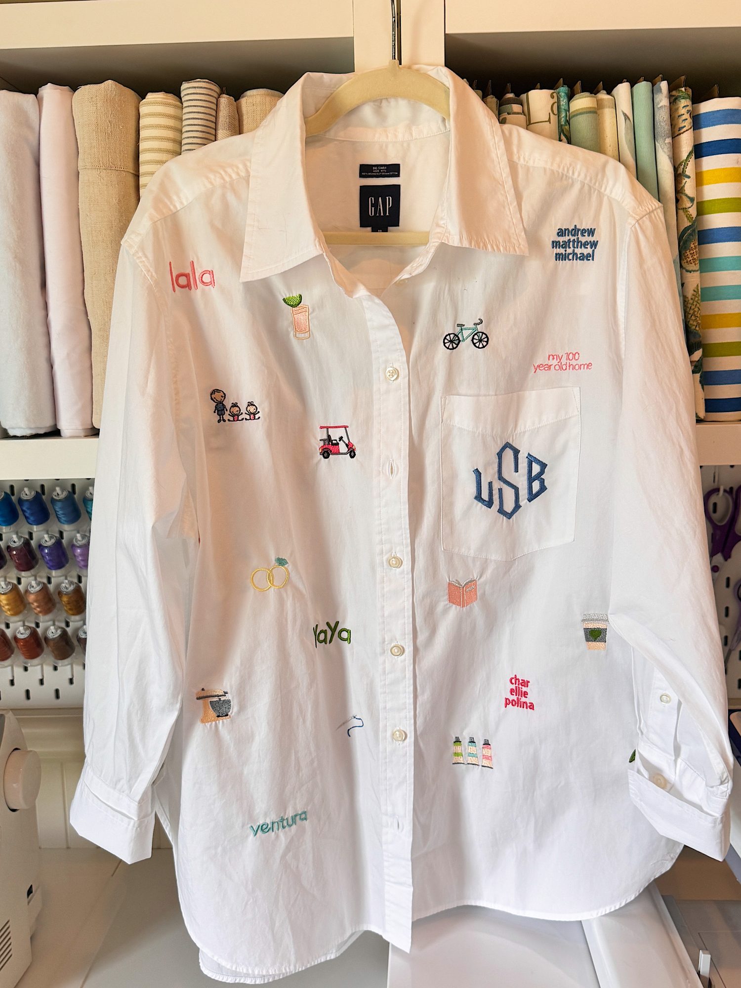 A white GAP button-up shirt hangs on a hanger, decorated with colorful embroidered words and small images. Sewing materials and fabric are visible in the background.