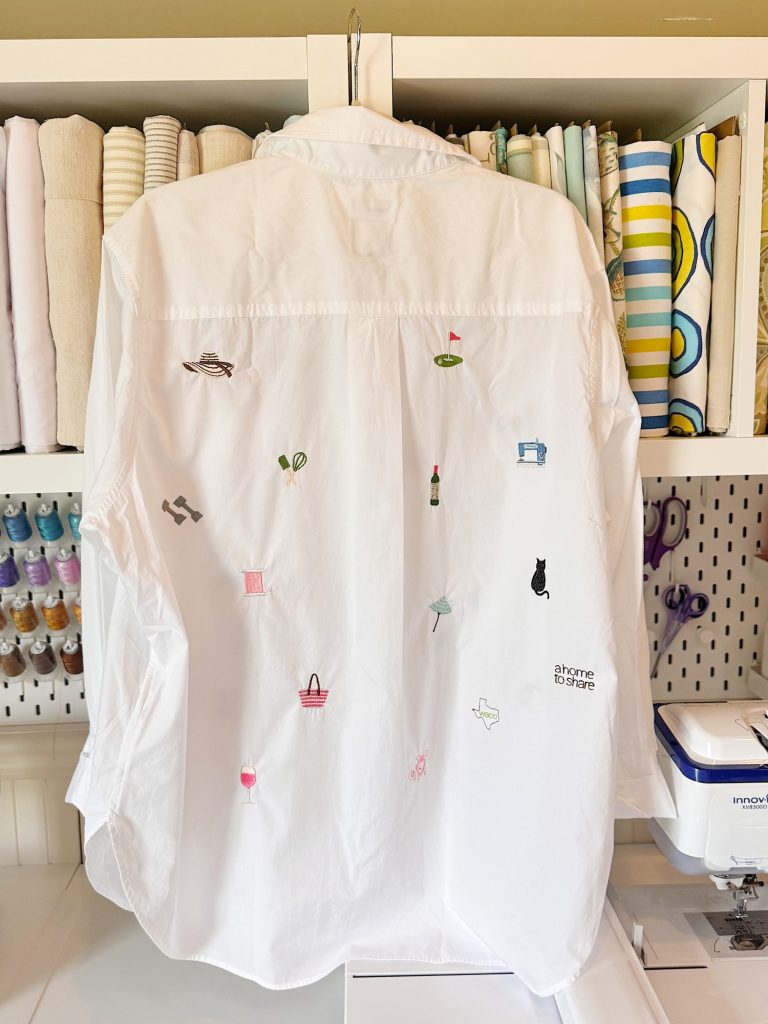 A white button-down shirt with small, colorful embroidered designs hangs in a sewing room, with folded fabrics and sewing supplies visible in the background.
